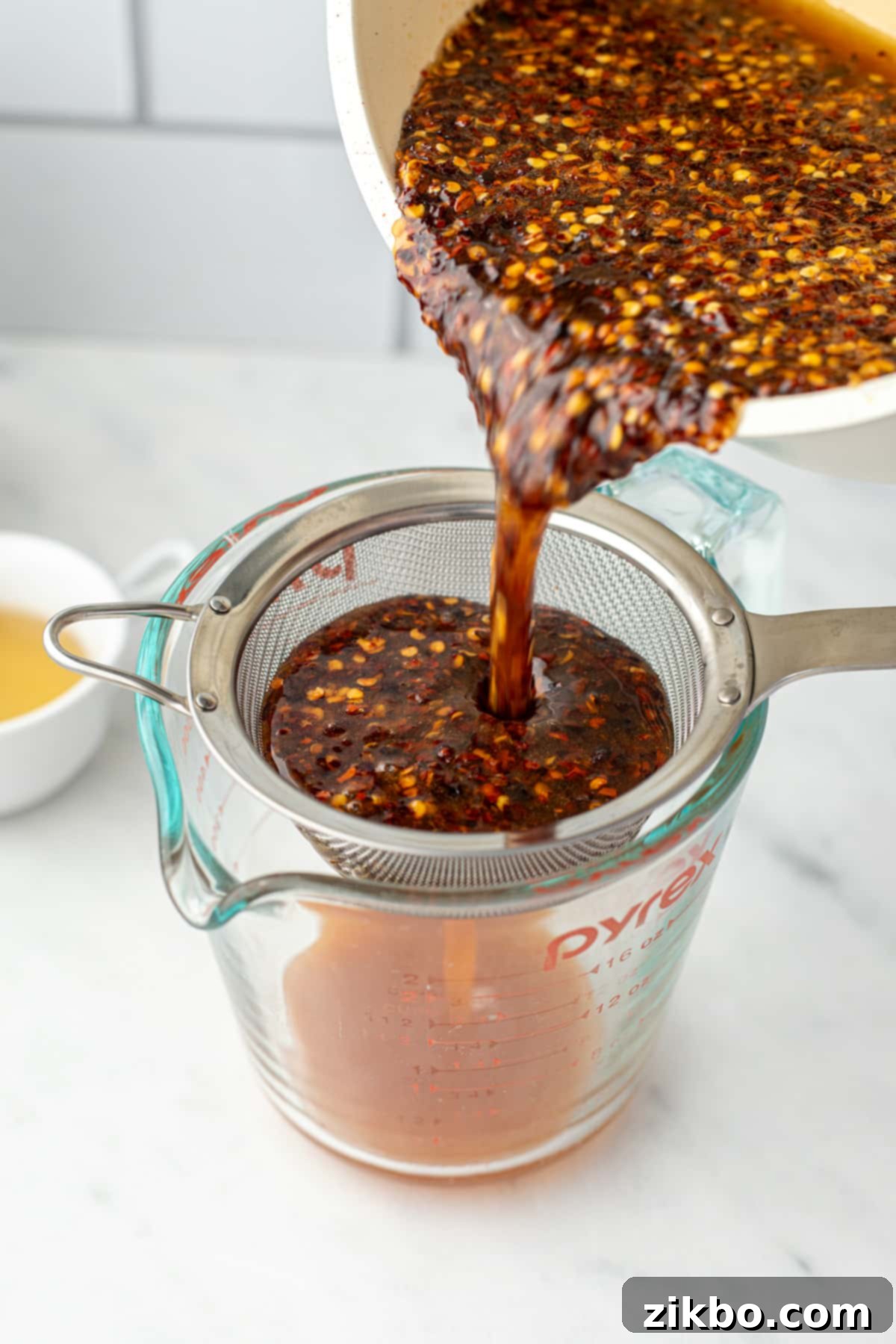 Carefully pouring the chili-infused honey through a fine mesh strainer into a glass measuring cup, separating the flakes.