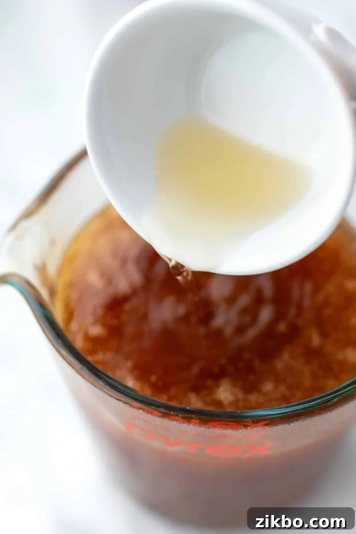 Pouring a stream of golden apple cider vinegar into a measuring cup filled with strained, infused hot honey.
