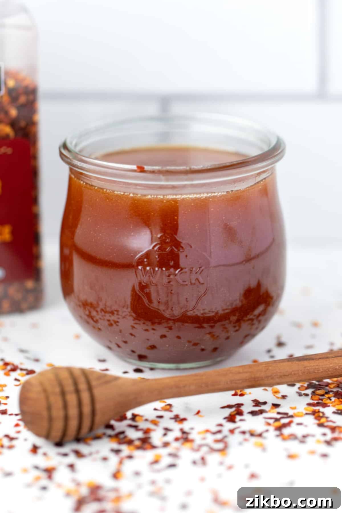A beautiful weck jar filled with golden hot honey, with a honey dipper resting on the counter beside scattered red pepper flakes, showcasing the final product.