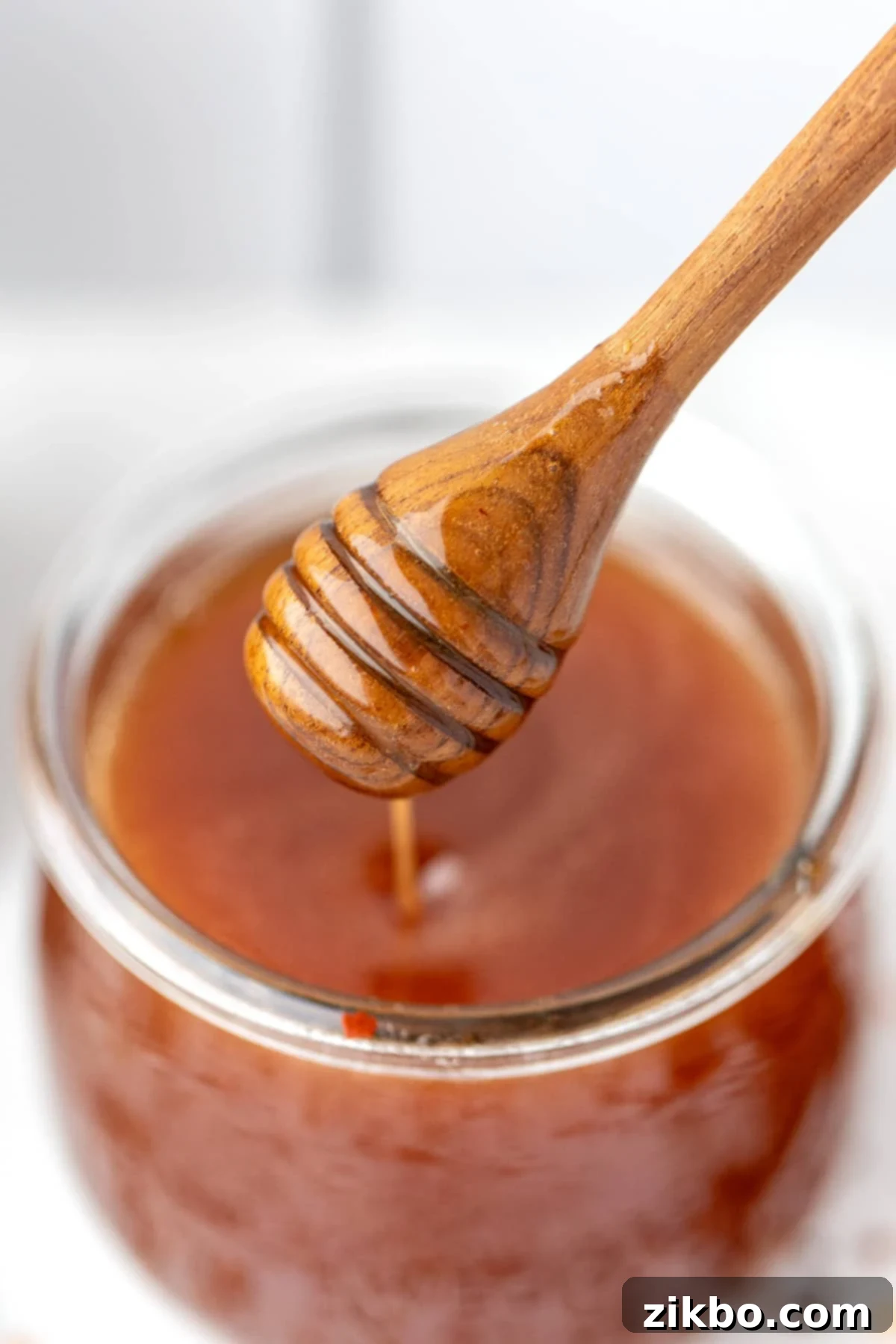A wooden honey dipper being pulled from a jar of golden hot honey, showing its rich, flowing texture.