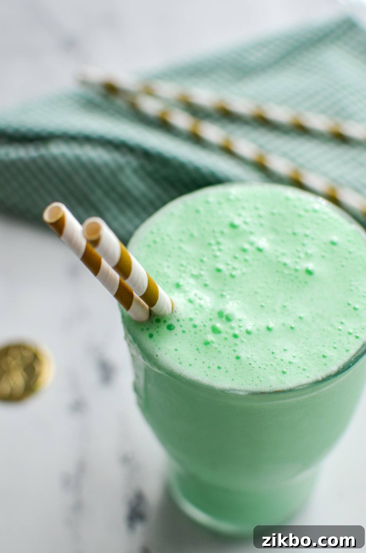 Homemade Shamrock Shake 2 A large glass holding a vibrant green milkshake, topped with whipped cream and two gold-striped straws, inviting a refreshing sip.