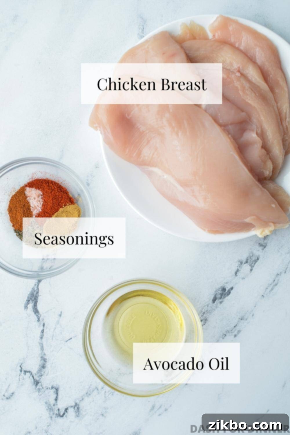 Raw chicken breasts, a bottle of avocado oil, and an assortment of aromatic seasonings arranged neatly on a cutting board, ready for preparation.