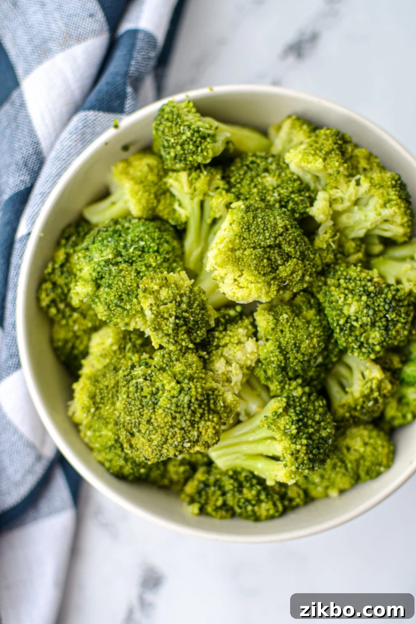 An overhead shot of a bowl of cooked broccoli, resting on a white marble counter and with a blue check napkin.