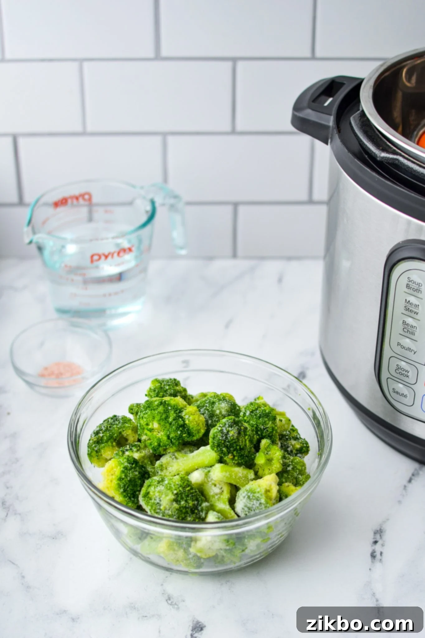 A bowl of frozen broccoli on a white marble countertop. A measuring cup of water, some salt, and an Instant Pot in the background.