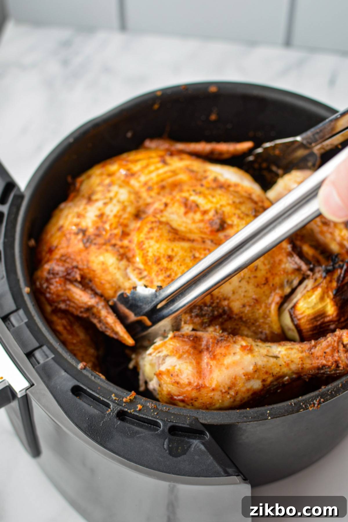 Using tongs to carefully flip a partially cooked whole chicken in an air fryer basket, ensuring the breast faces up.
