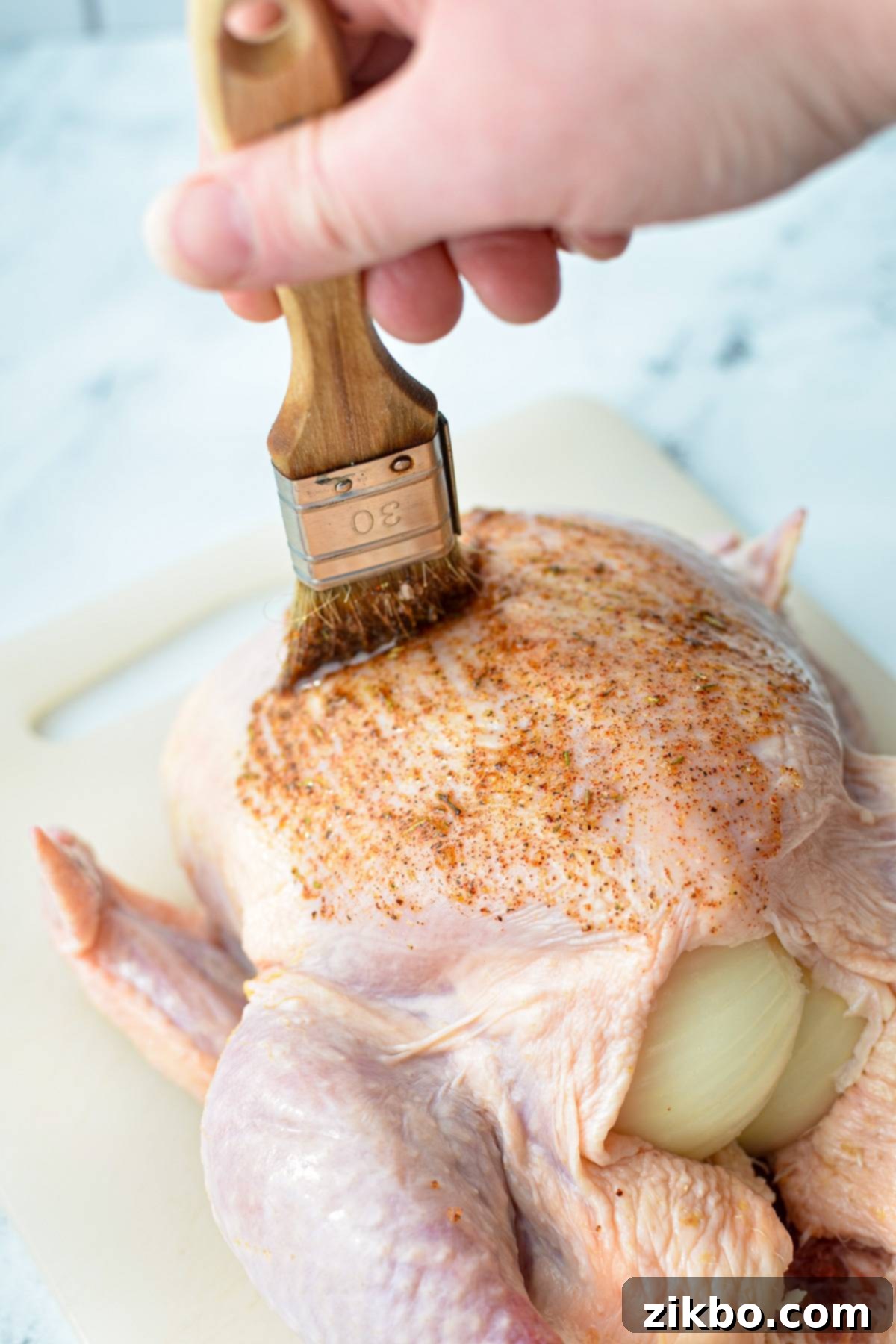 A raw whole chicken being brushed with a homemade seasoning blend on a cutting board.