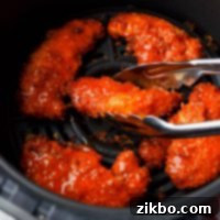 An air fryer basket filled with buffalo chicken tenders, and a pair of tongs taking them out.