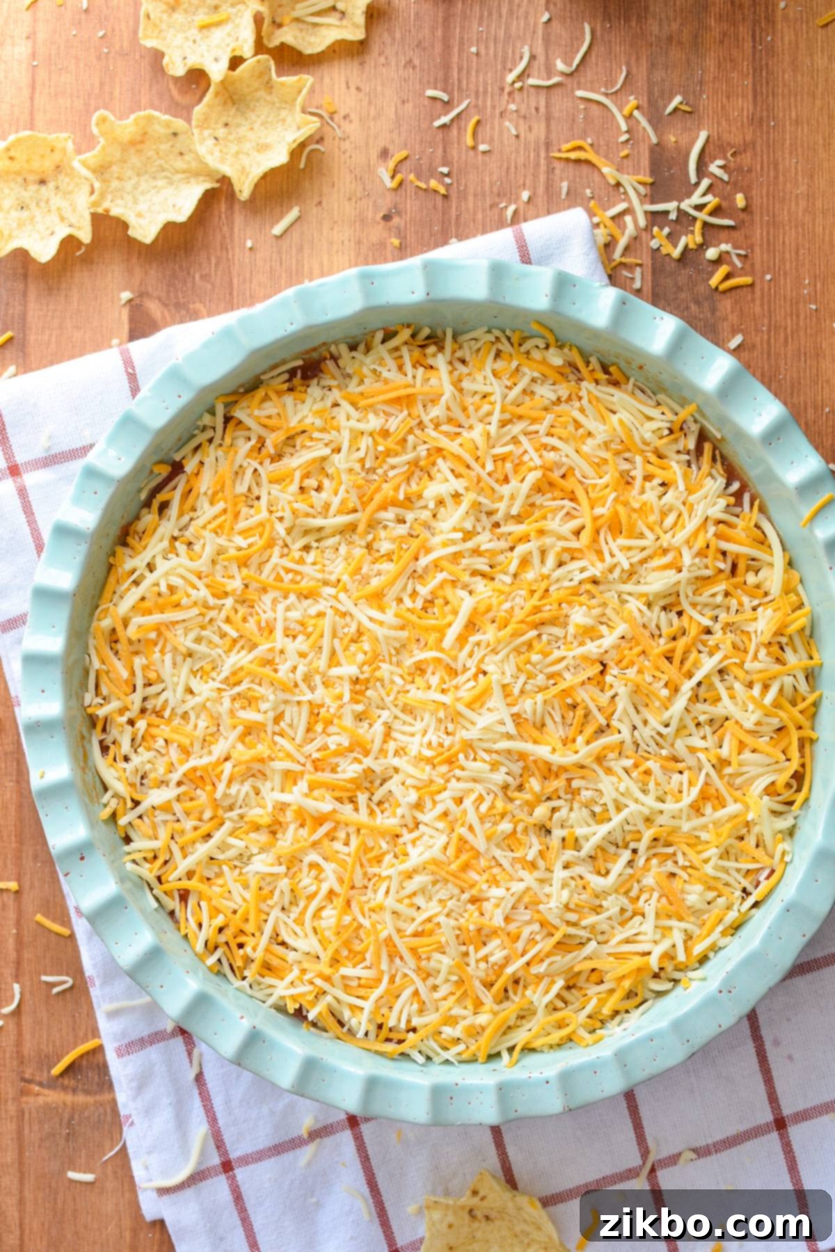 A perfectly crafted circular layered nacho dip in a white dish, adorned with a generous topping of golden shredded cheese, showcasing its inviting and delicious layers.