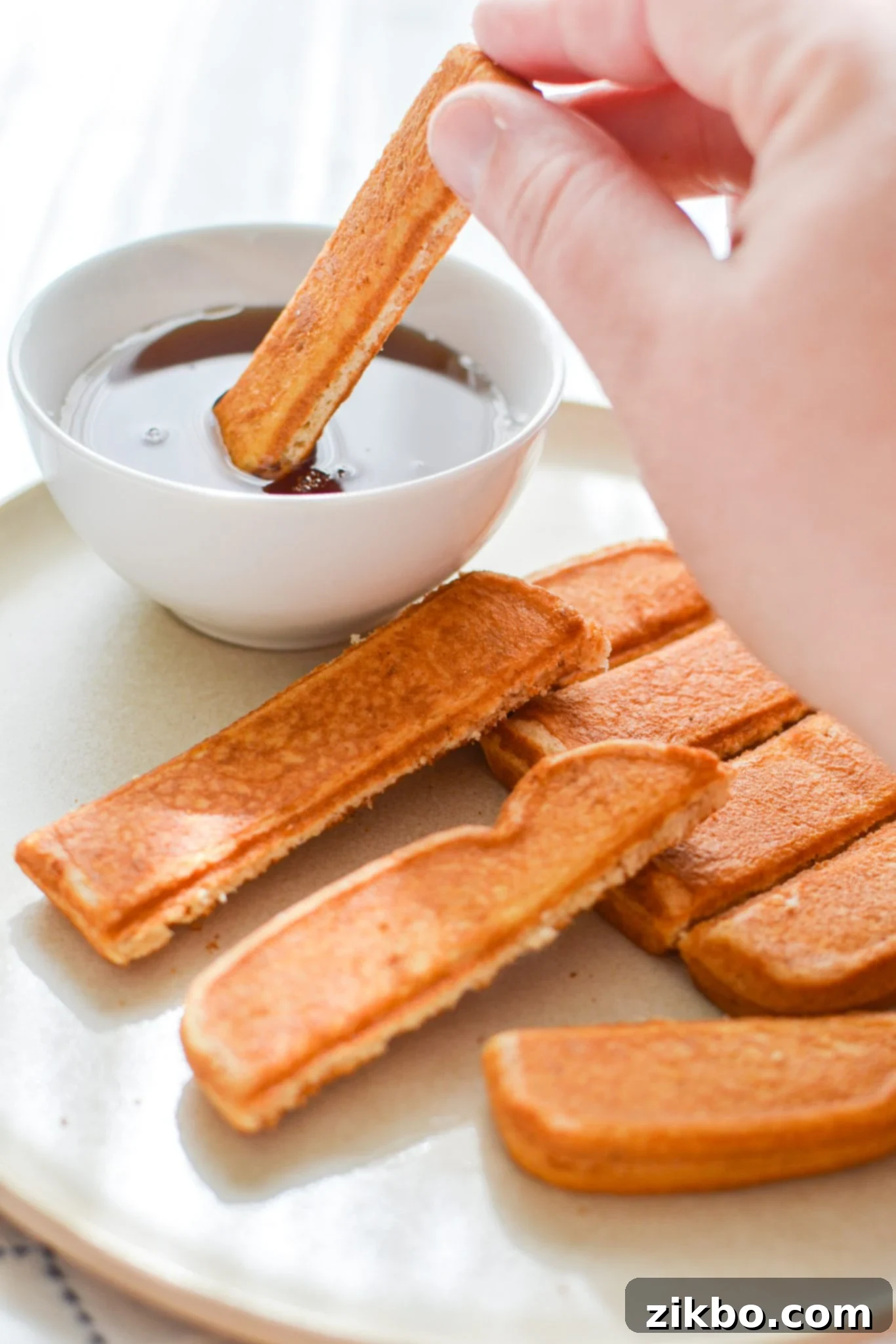Close-up of a perfectly cooked, crispy French toast stick being dipped into a bowl of rich maple syrup.