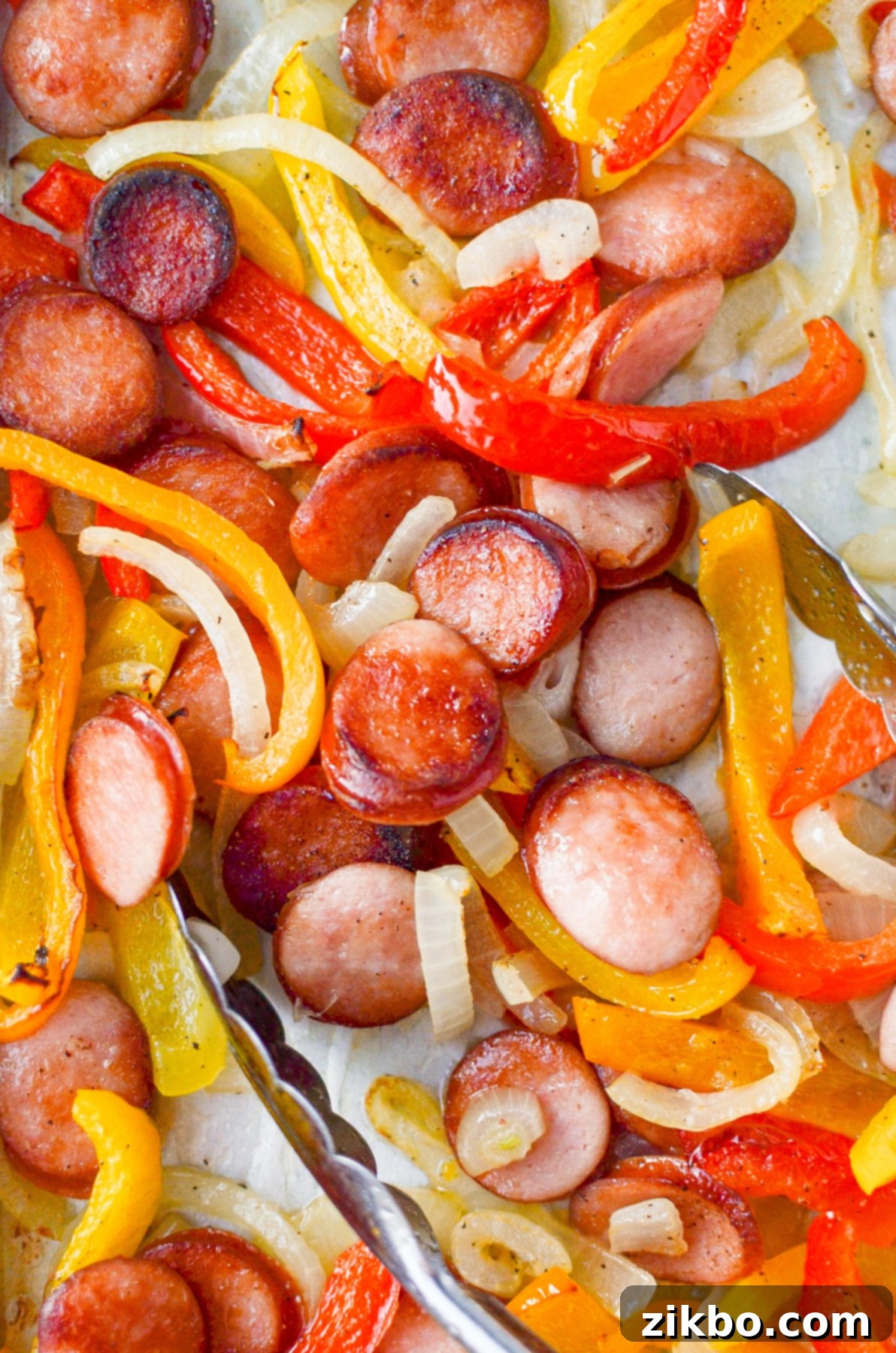 A pair of tongs grabbing some food from a sheet pan, showcasing the delicious cooked sausage and peppers.