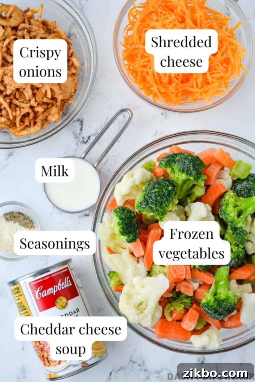 A flat lay of ingredients for cheesy vegetable casserole: a bag of frozen mixed vegetables, condensed cheese soup, milk, shredded cheddar cheese, and a container of crispy fried onions.