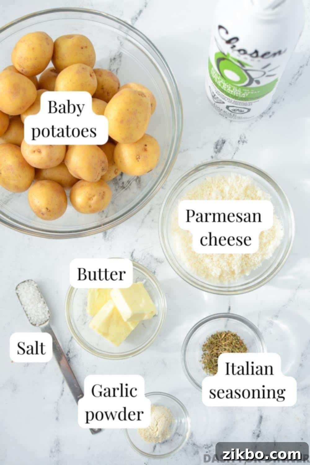 Crispy Parmesan Potatoes 3 The ingredients needed to make parmesan crusted baby potatoes: baby potatoes, parmesan cheese, butter, Italian seasoning, garlic powder, salt, and cooking spray laid out on a wooden surface.