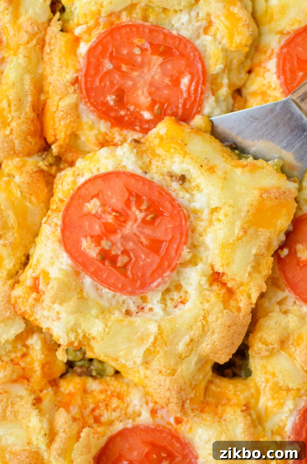 A spatula lifting a cheesy casserole from a dish topped with fresh tomato slices.