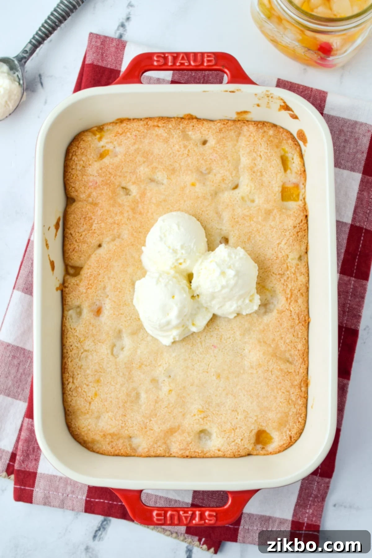 A vibrant red baking dish filled with a golden cake, elegantly topped with three scoops of creamy vanilla ice cream, ready to be served.