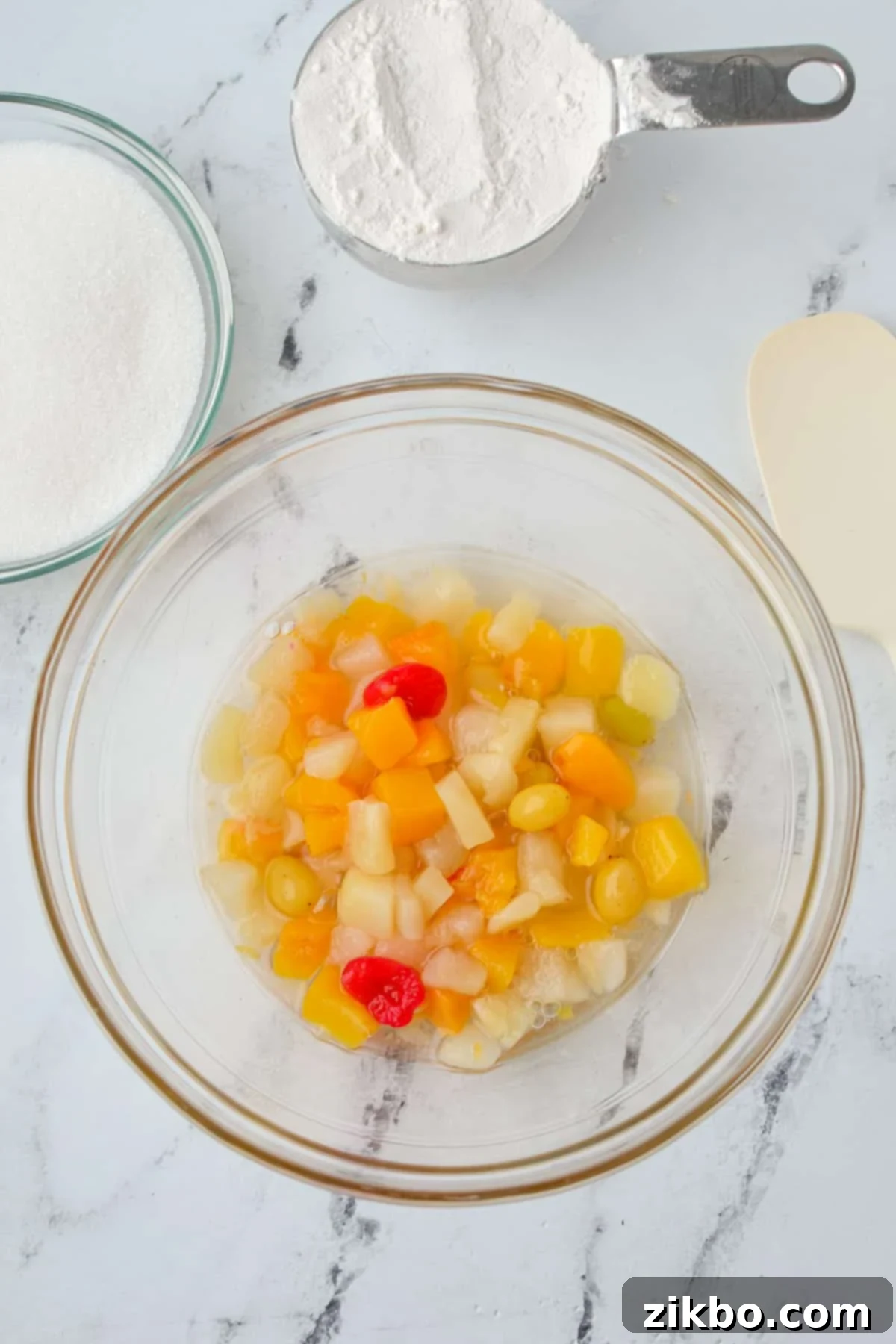 A medium-sized glass mixing bowl holds a cup of colorful fruit cocktail, including its luscious juice, ready for the next ingredients.