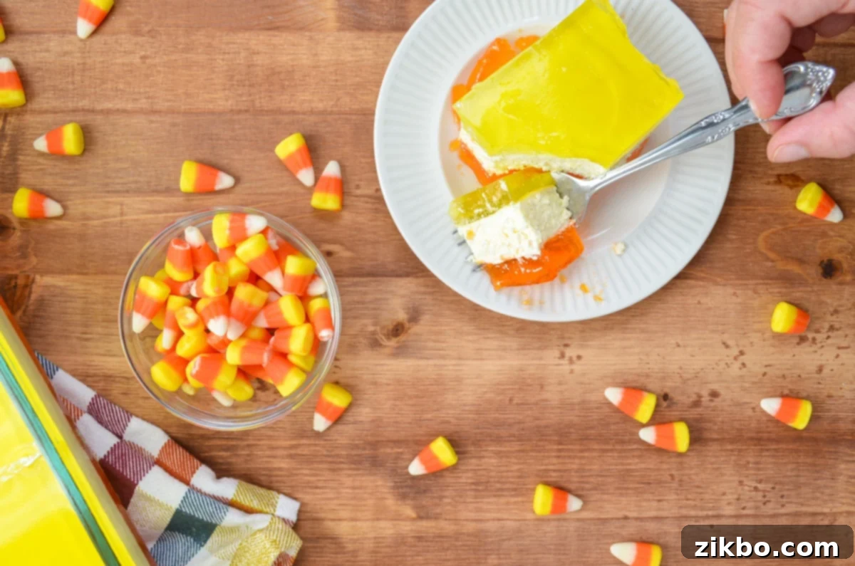 A vibrant layered lemon and orange Jell-O dessert being cut into with a fork, with a bowl of classic candy corn beside it.