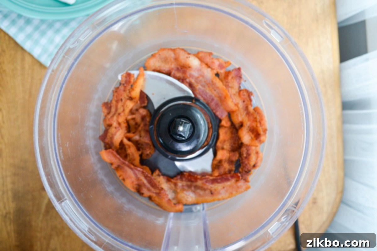 Cooked bacon slices being added to the bowl of a food processor to make bacon bits.
