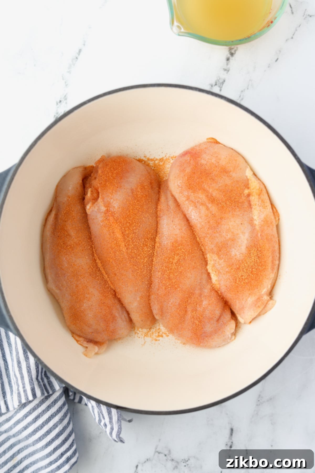 A Dutch Oven with four raw chicken breasts, generously seasoned with salt and spices.