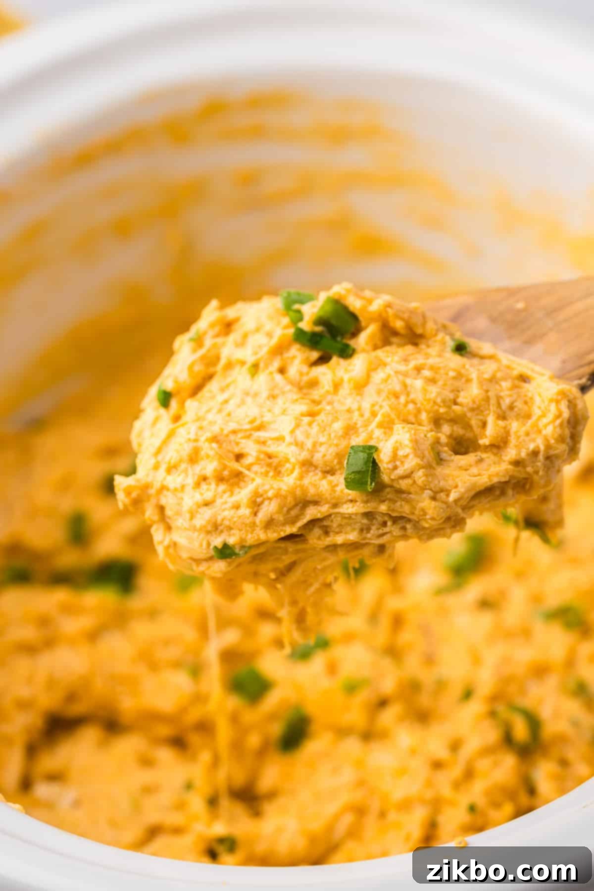 A wooden spoon scooping a generous portion of the hot, creamy buffalo chicken dip from a white slow cooker, showing its inviting texture.