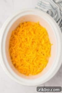A white crock pot with a cheesy dip inside, topped with shredded cheddar cheese.