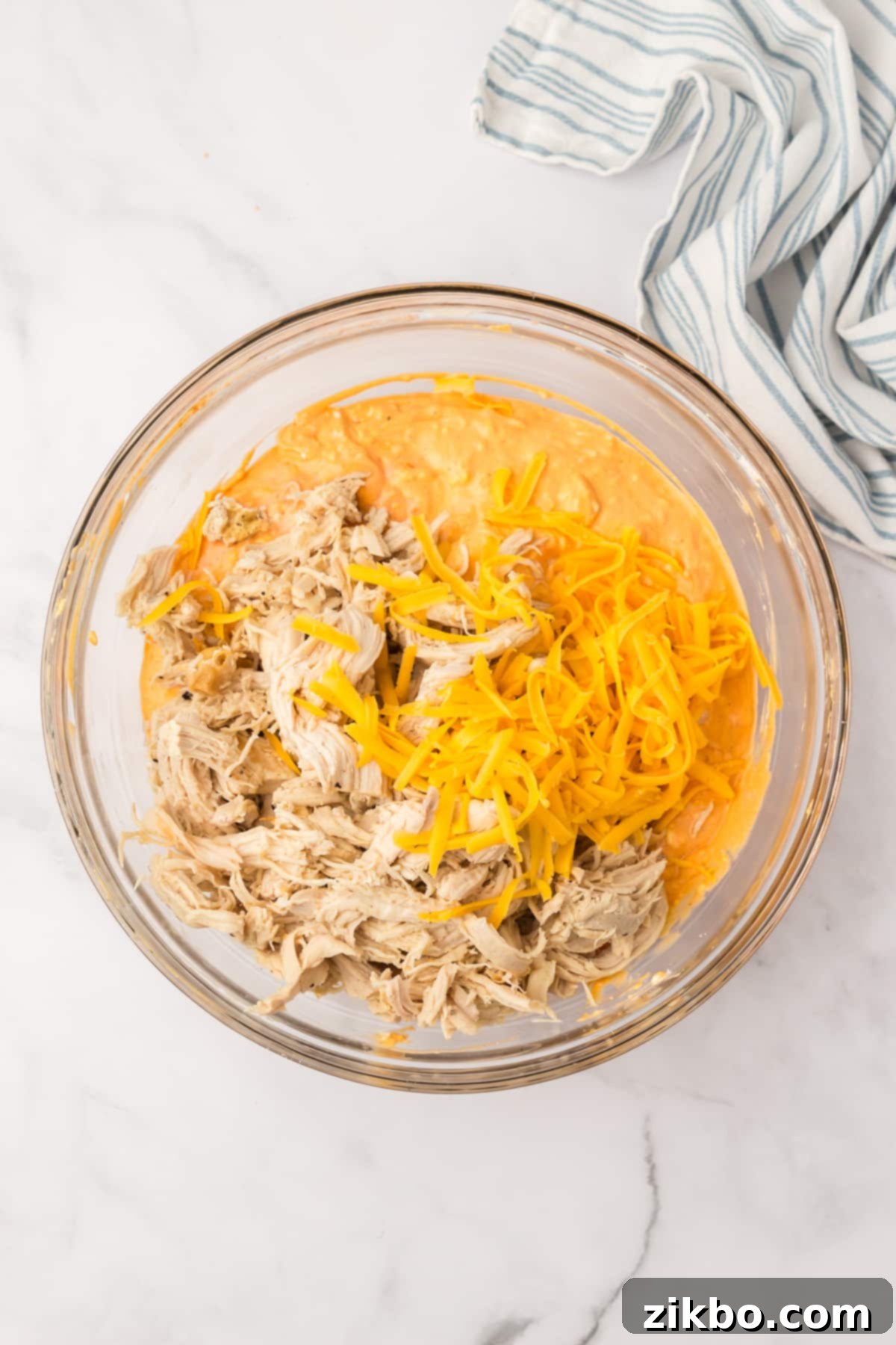 A bowl filled with the creamy dip mixture, with shredded cooked chicken and a portion of shredded cheddar cheese layered on top, ready to be mixed.