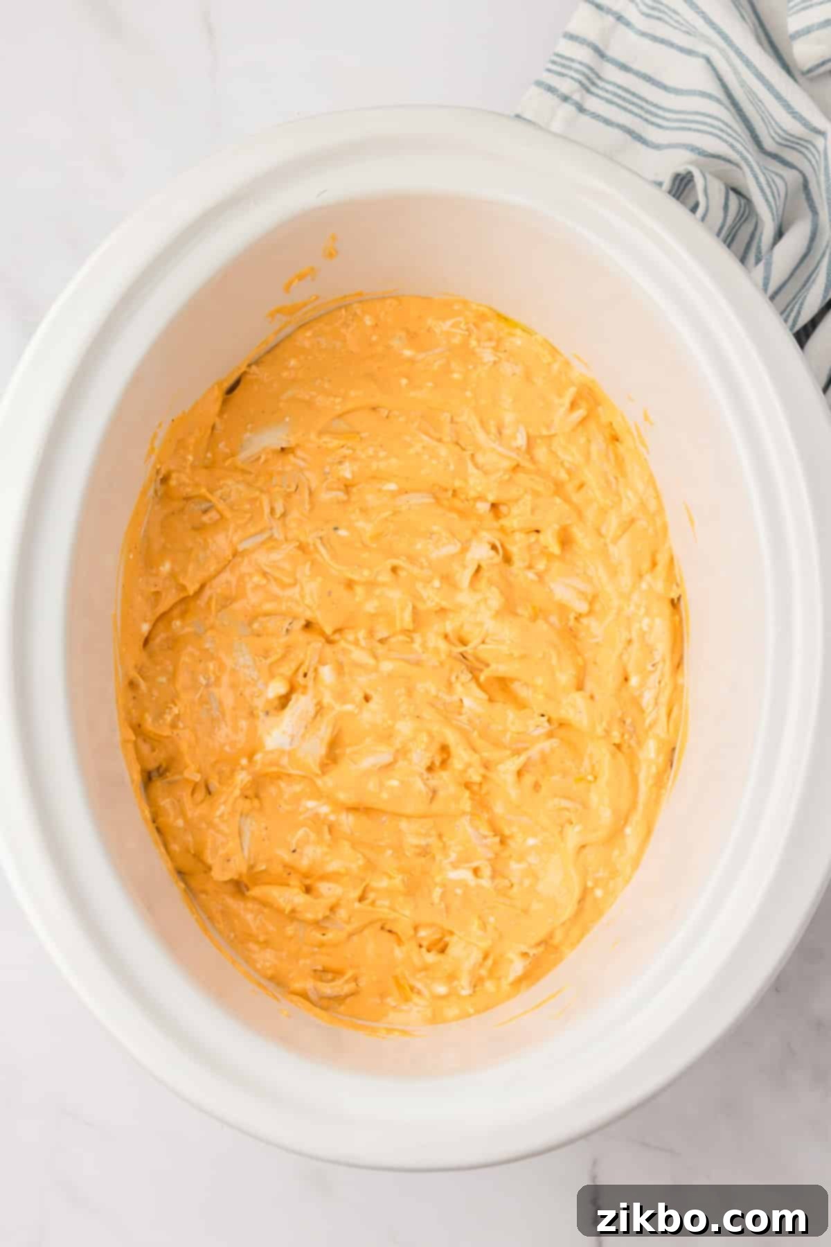 A white slow cooker pot filled with the unheated buffalo chicken dip mixture, spread evenly at the bottom.