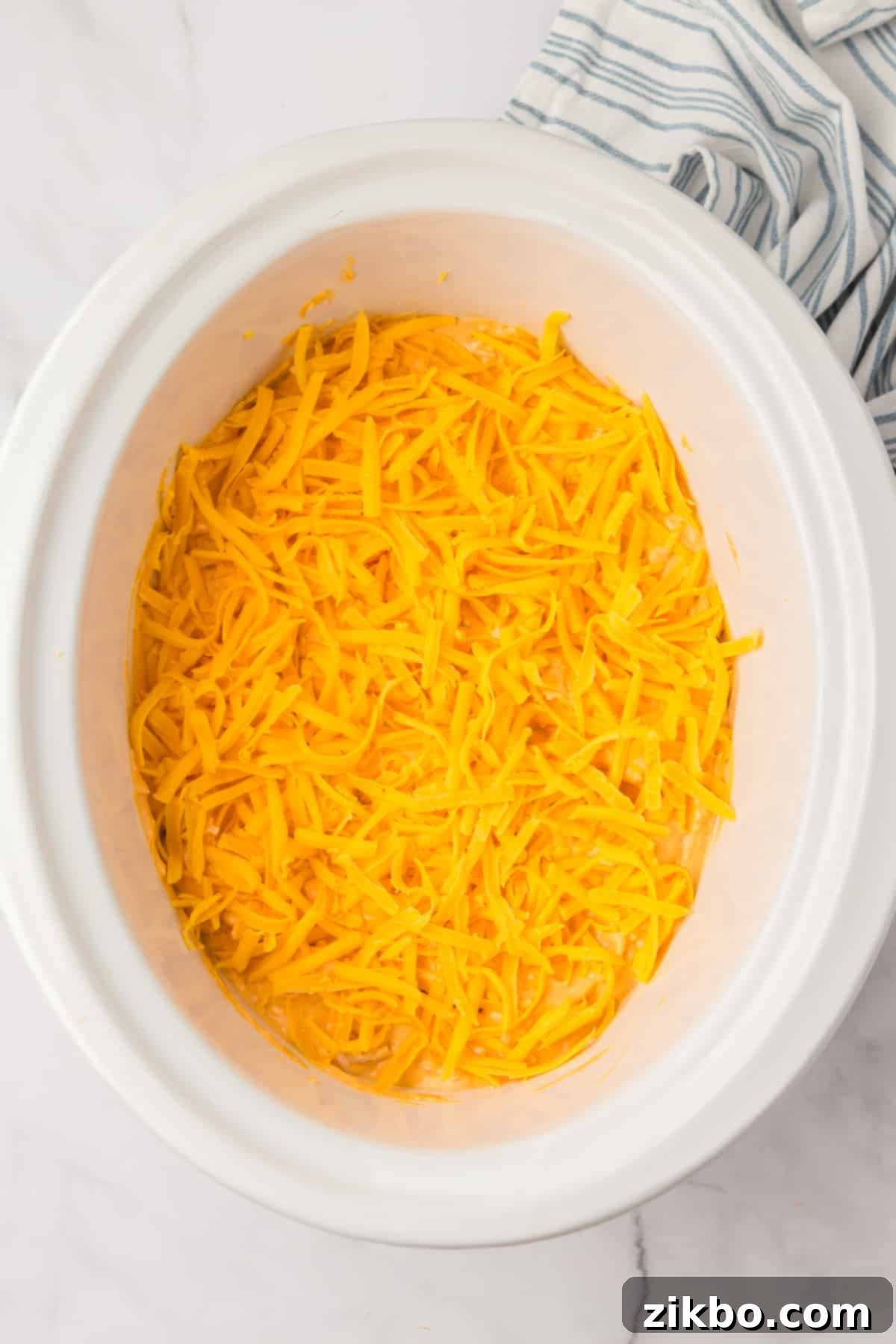 A white slow cooker pot with the buffalo chicken dip, now topped with the remaining shredded cheddar cheese, ready for cooking.