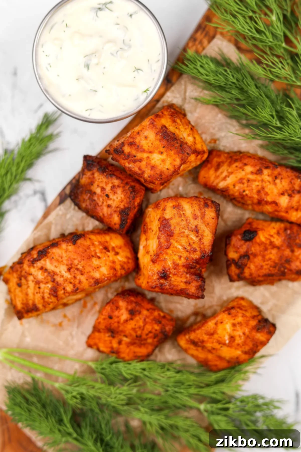 Cooked salmon bites arranged neatly on a plate with a small ceramic dish of creamy dill sauce beside them.