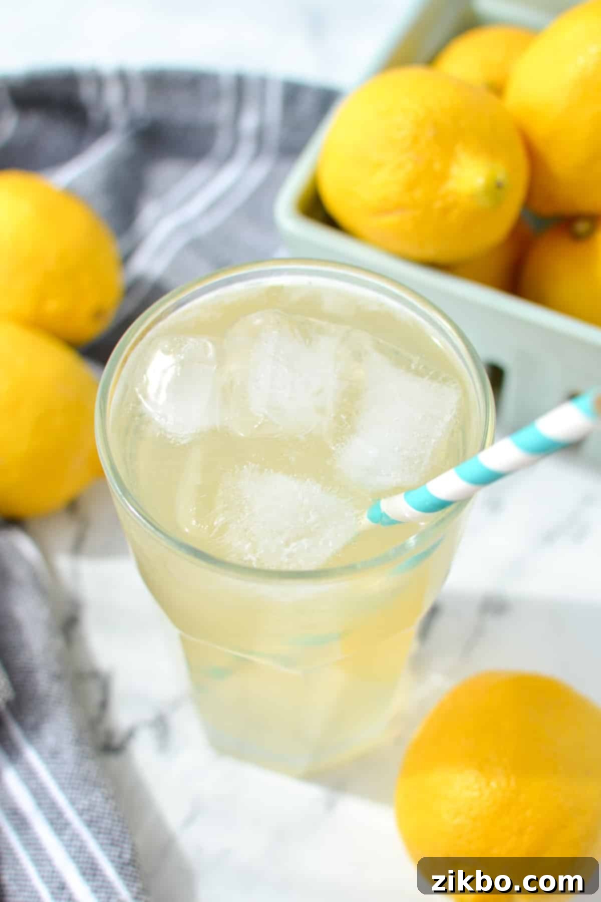 Lemonade in a glass filled with ice, with fresh lemons and a mint sprig in the background.