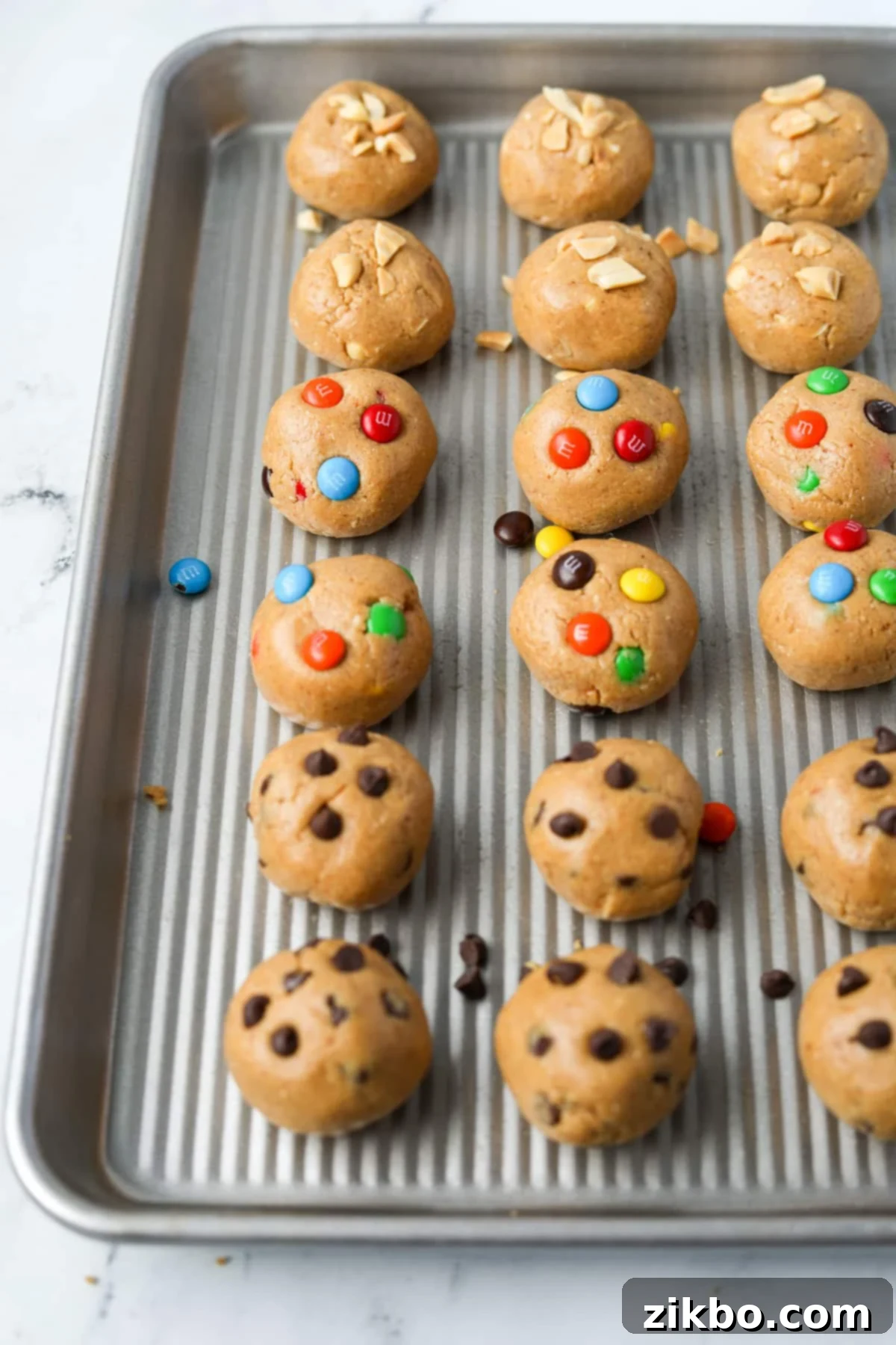 A baking tray with cookie dough balls, featuring chocolate chip, m&m, and chopped peanuts.