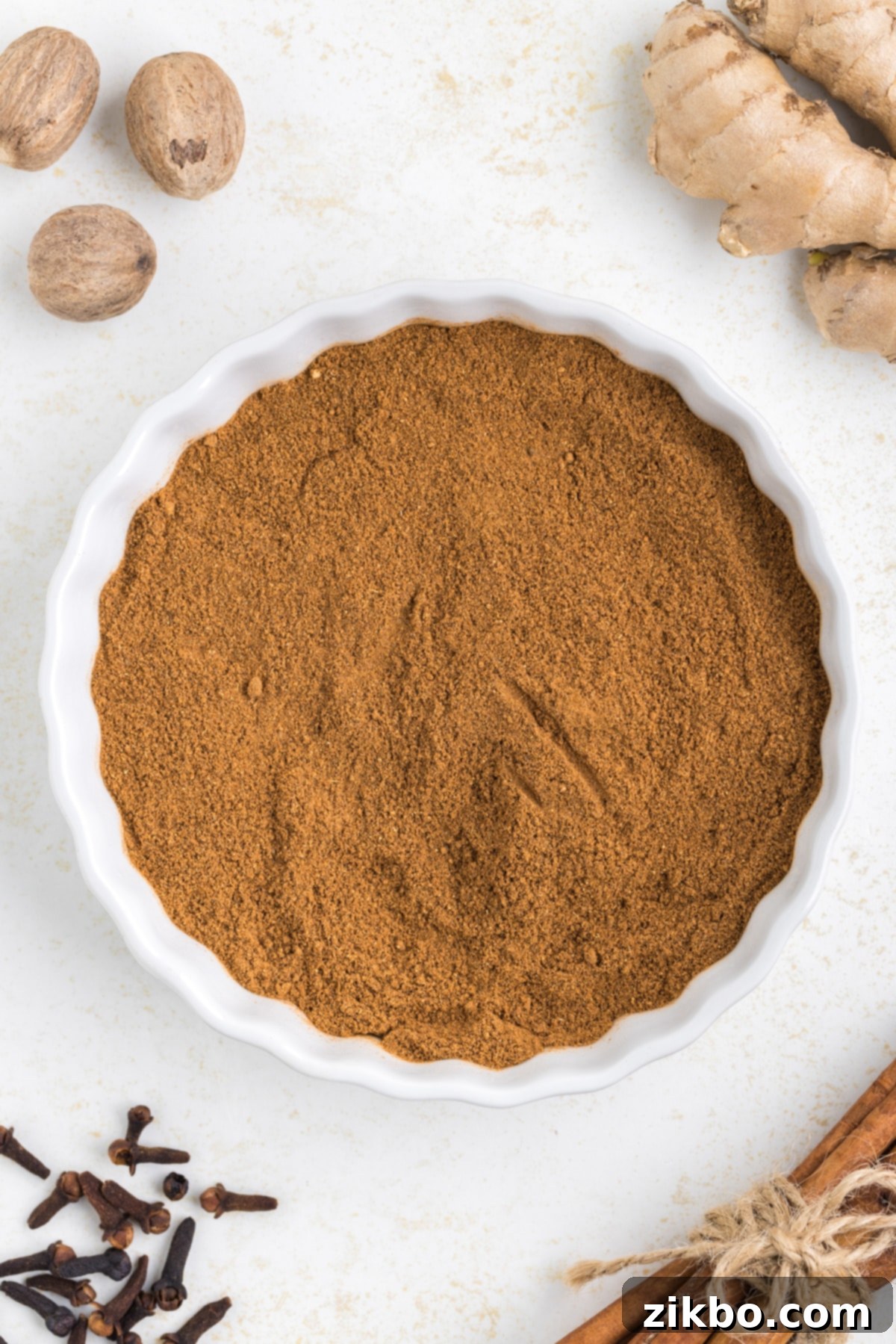 A dish filled with a newly mixed homemade spice blend, surrounded by whole ginger root, nutmeg, cloves, and cinnamon sticks.