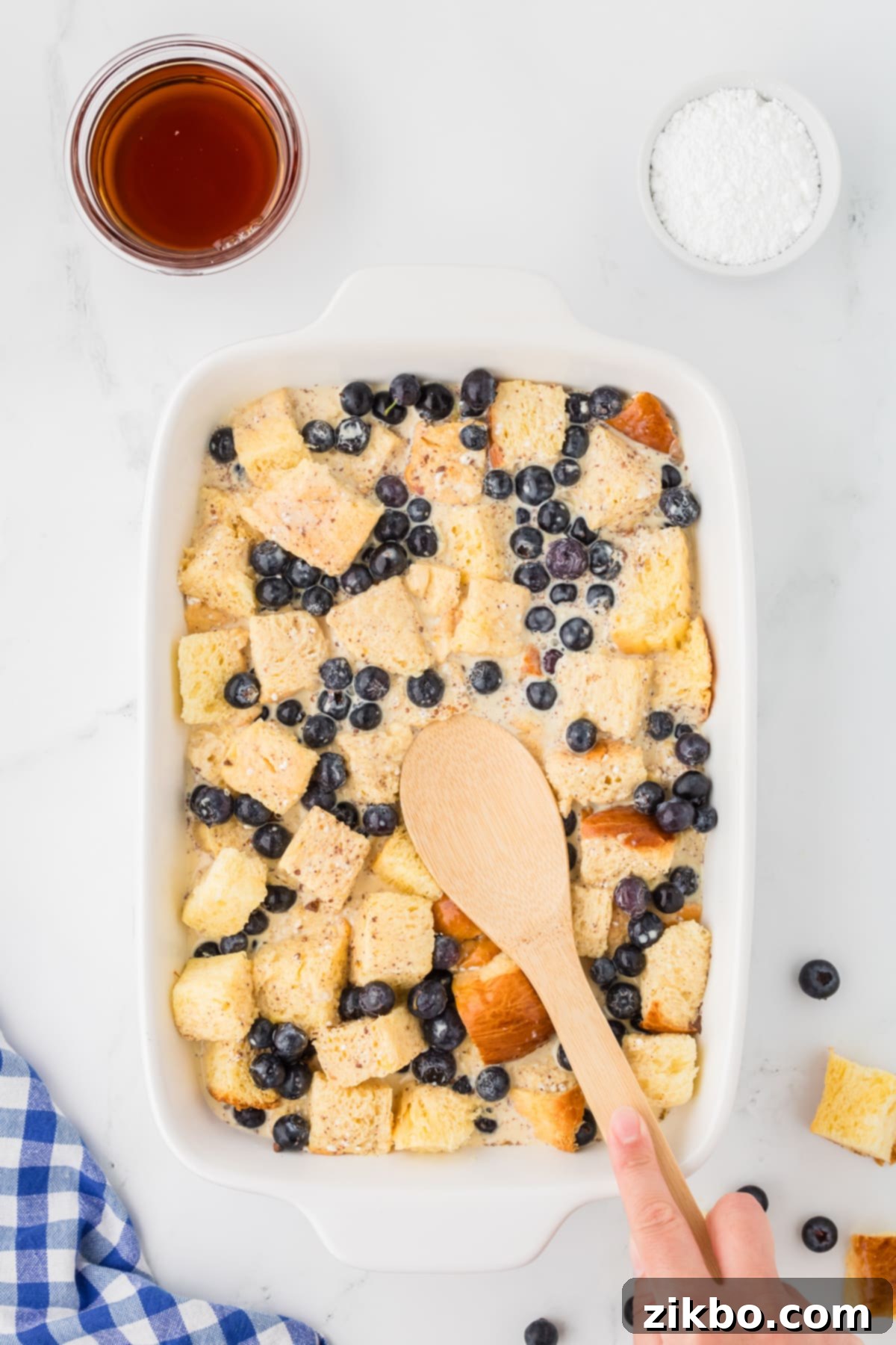 Overnight Brioche French Toast Bake 8 Using a wooden spoon to press bread cubes into a custard mixture in a baking dish.