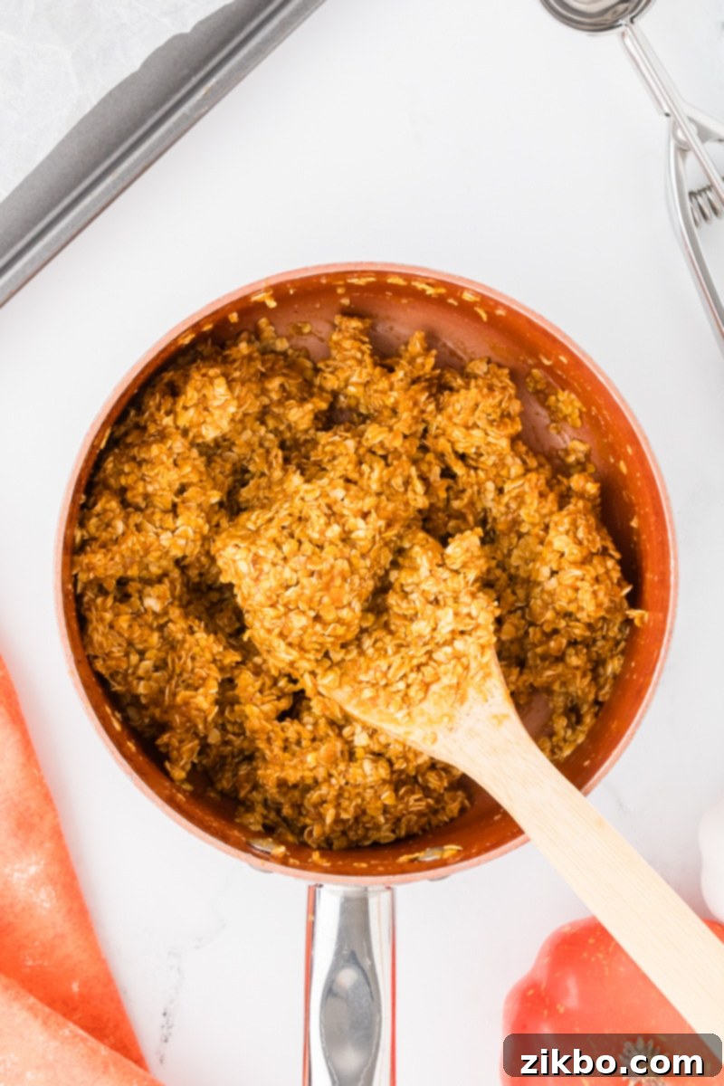 Spiced Pumpkin No-Bake Bites 7 An orange pumpkin mixture in a saucepan being thoroughly mixed with quick oats using a wooden spoon until fully coated.