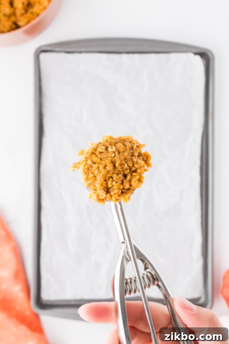 Spiced Pumpkin No-Bake Bites 8 A cookie scoop holds a portion of the pumpkin oatmeal mixture, poised over a parchment-lined baking sheet.