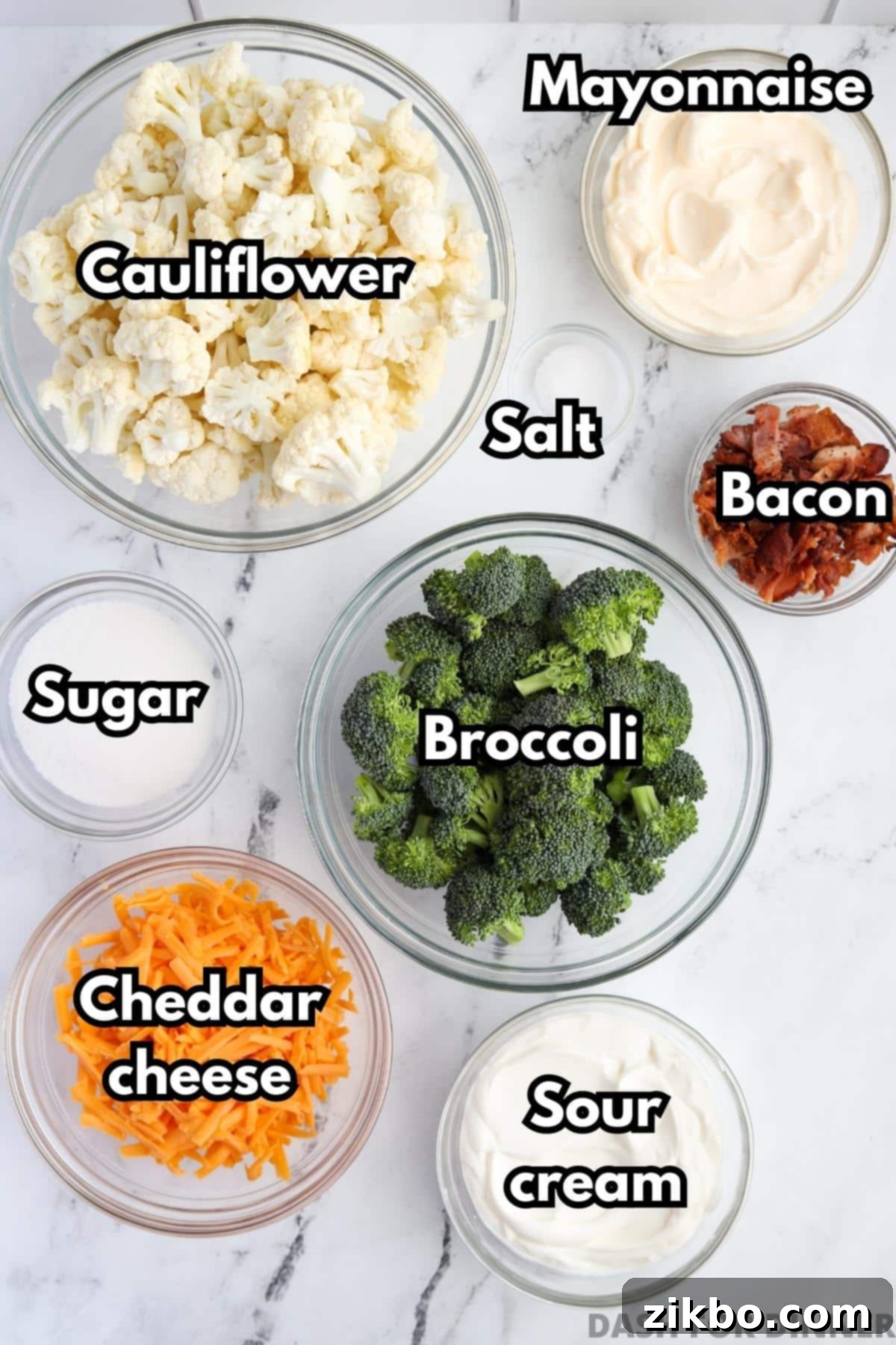 A flat lay image displaying all the fresh ingredients needed for a delicious broccoli salad: fresh broccoli and cauliflower florets, crispy bacon, shredded sharp cheddar cheese, mayonnaise, sour cream, sugar, and a pinch of salt.