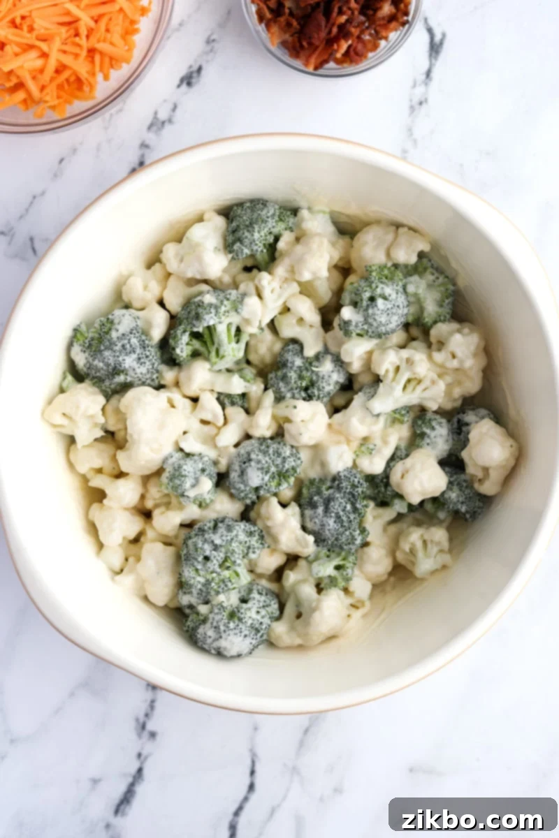 Raw broccoli and cauliflower florets thoroughly coated in a thick, creamy white dressing in a large mixing bowl.