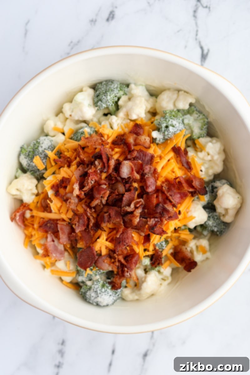 A bowl of creamy broccoli and cauliflower salad, with a generous sprinkle of shredded cheddar cheese and crumbled crispy bacon added on top.