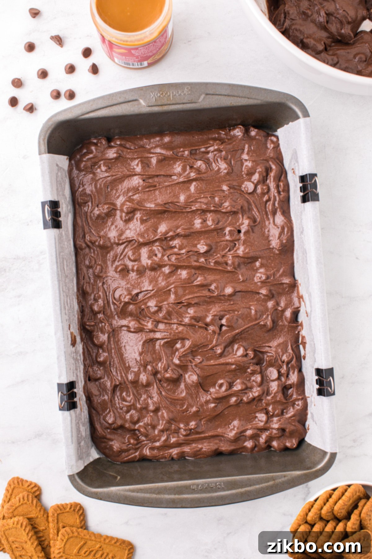 A 9x13 inch baking pan, neatly lined with parchment paper and containing an even base layer of rich brownie batter.