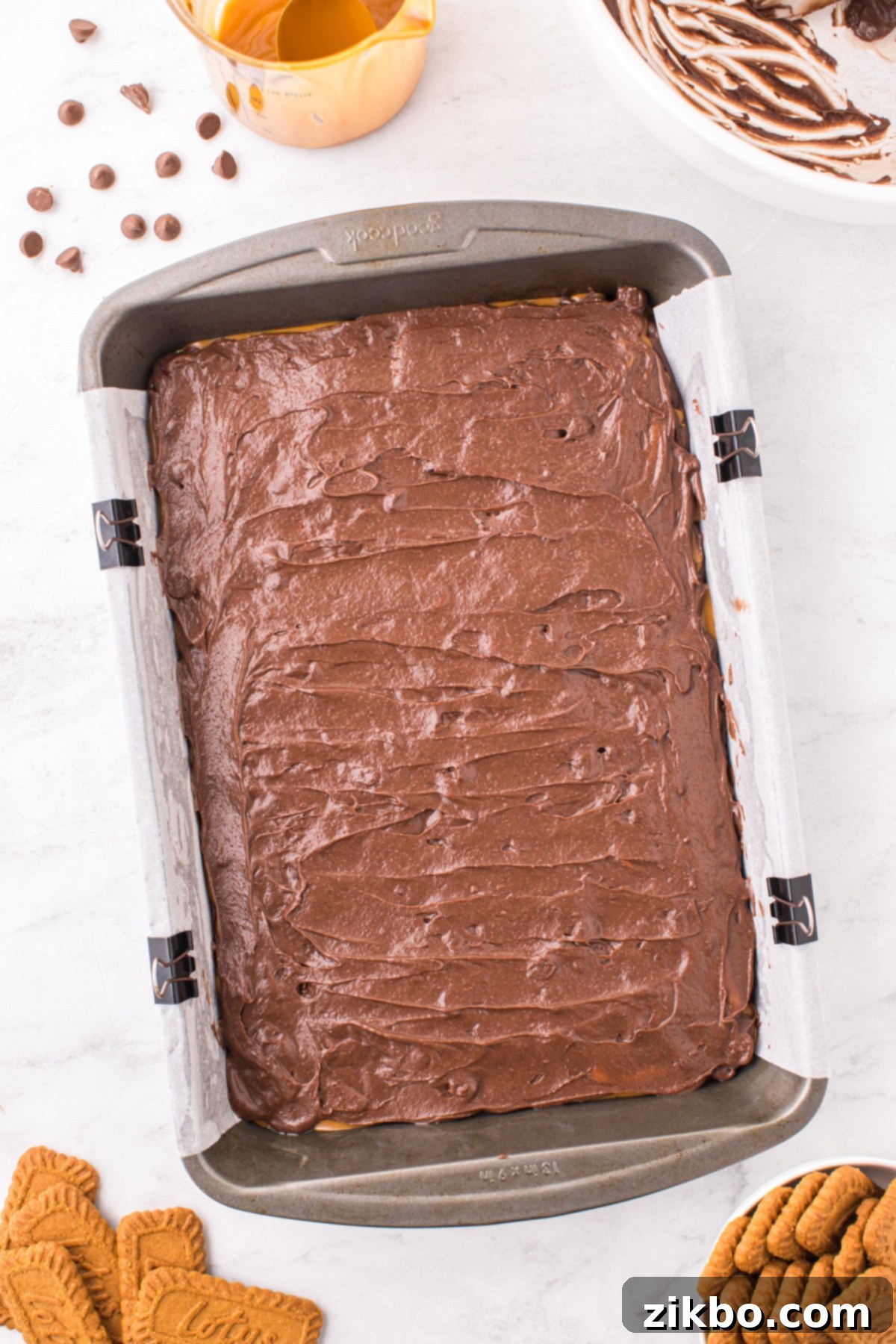 An unbaked brownie dish with a base layer of brownie batter, a middle layer of Biscoff spread, and the final layer of brownie batter gently spread on top, ready for the crushed cookies.