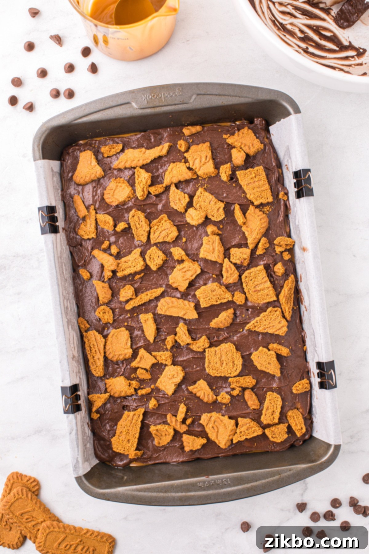 A baking pan filled with unbaked Biscoff brownie batter, generously topped with roughly chopped Lotus Biscoff cookie pieces, ready for the oven.