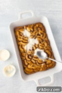 Spreading glaze on a baking dish with baked cinnamon roll pieces.
