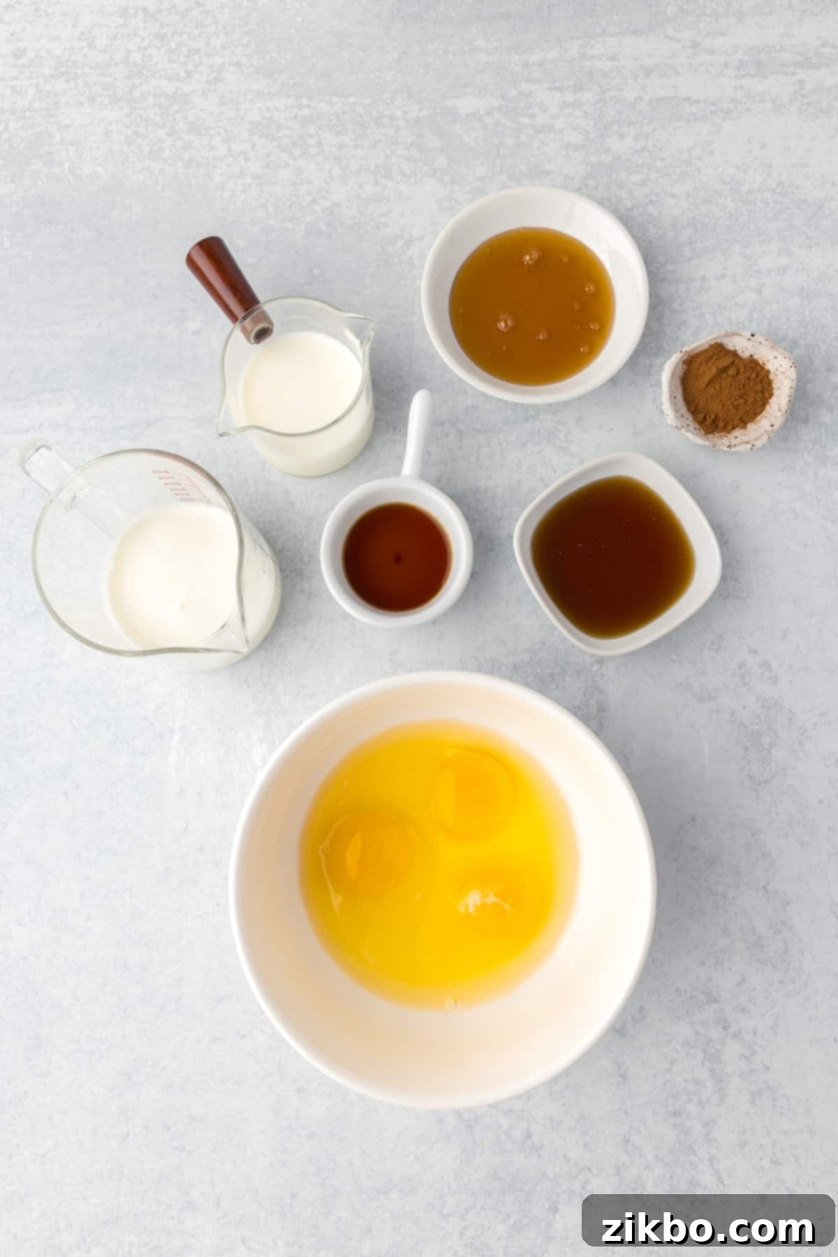 A bowl of whisked eggs, with small cups and bowls of other custard ingredients arranged around it on a table.