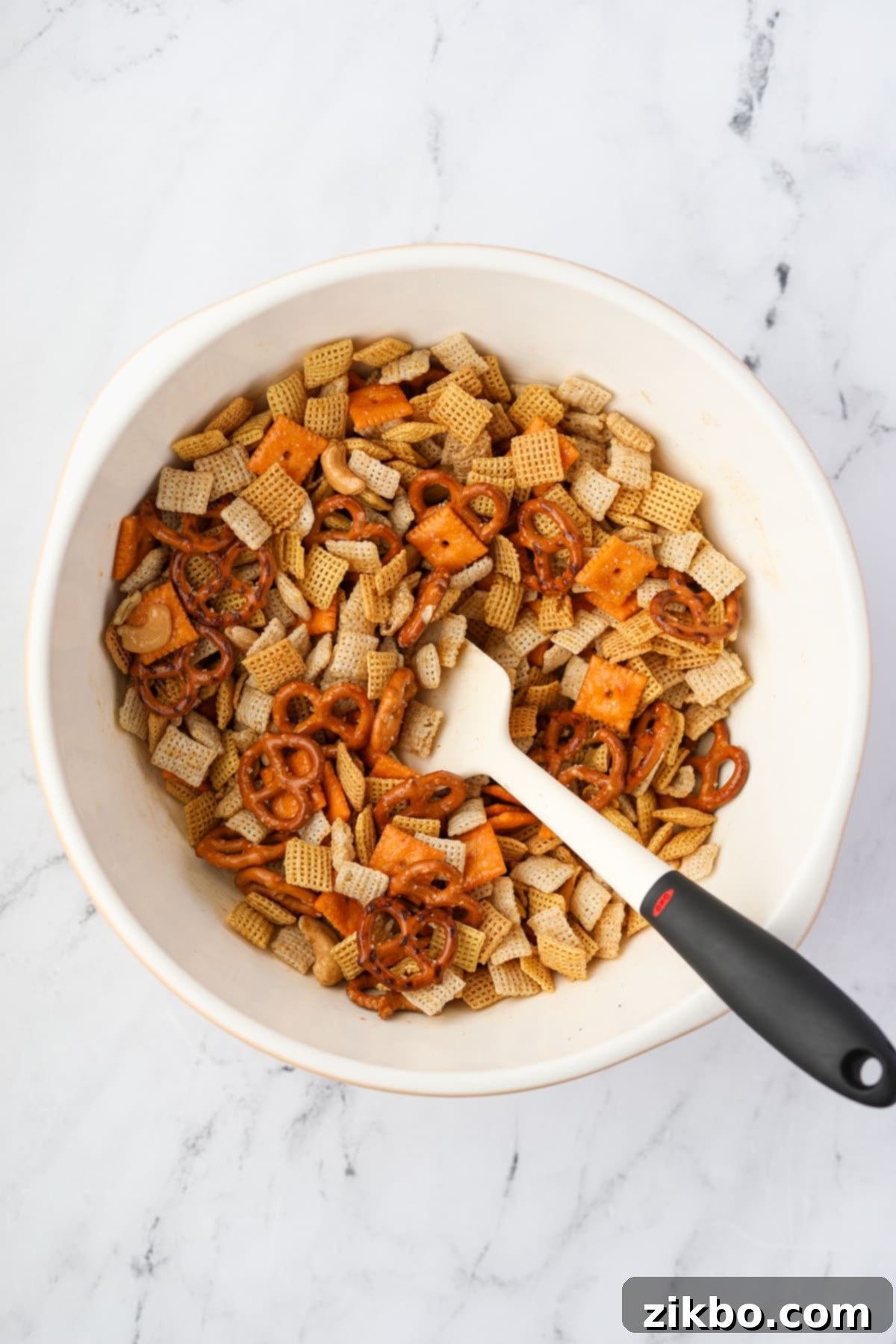A bowl of chex mix being stirred with a silicone spatula.