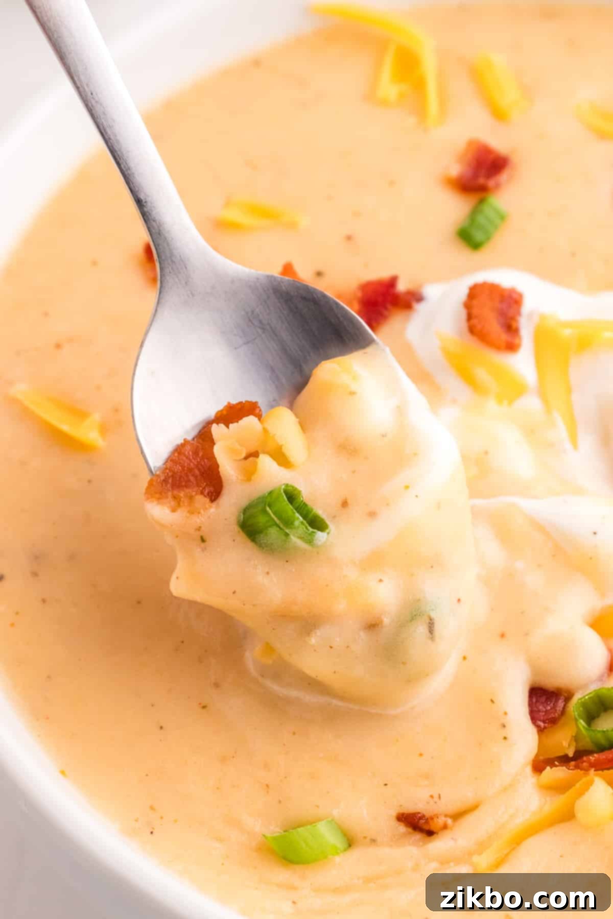 A spoon lifting a creamy portion of baked potato soup, adorned with fresh green onions and crispy bacon bits, from a bowl.