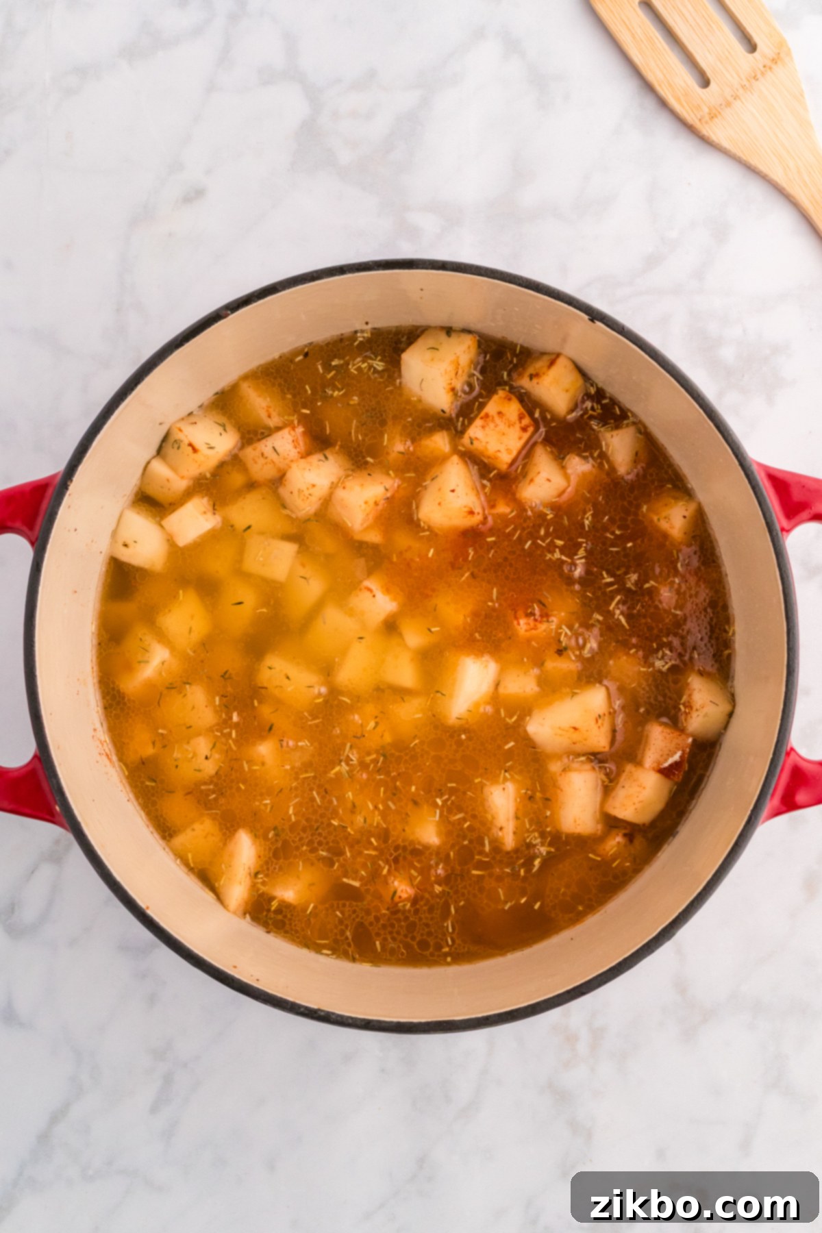 Chicken broth being poured over seasoned potatoes and onions in a Dutch oven, ready for simmering.