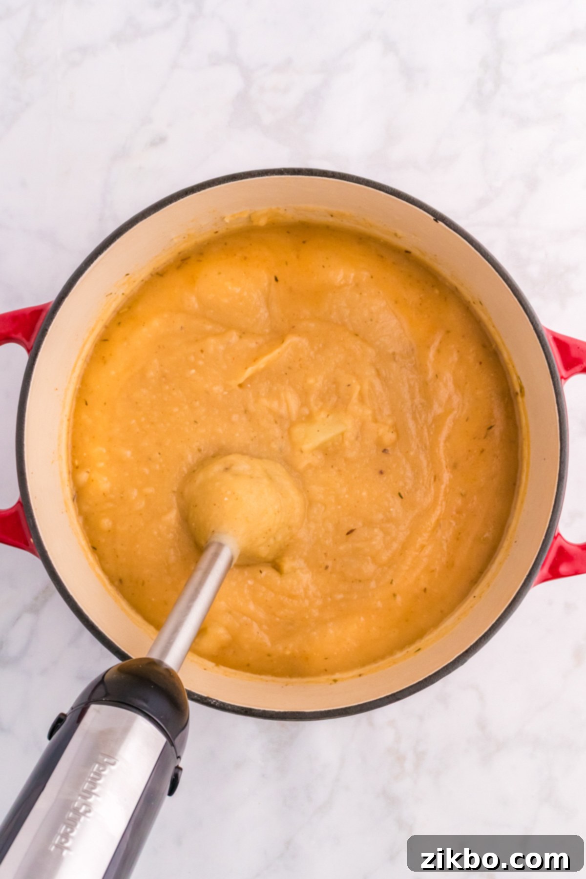 An immersion blender being used to puree cooked potatoes in a Dutch oven, creating a creamy soup base while leaving some chunks.