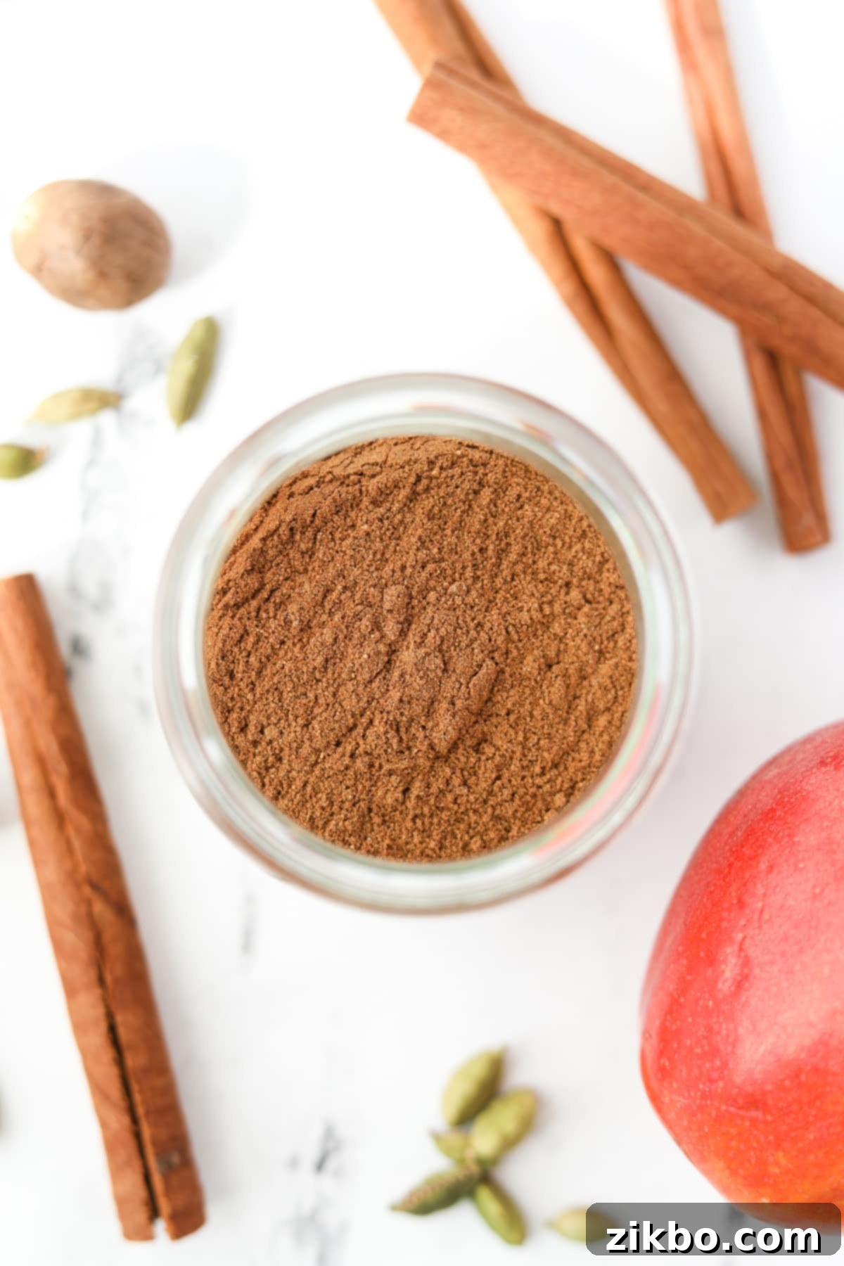 A small jar of apple spice on a marble counter, surrounded by cinnamon sticks, cloves, nutmeg, and cardamom pods, emphasizing natural ingredients.