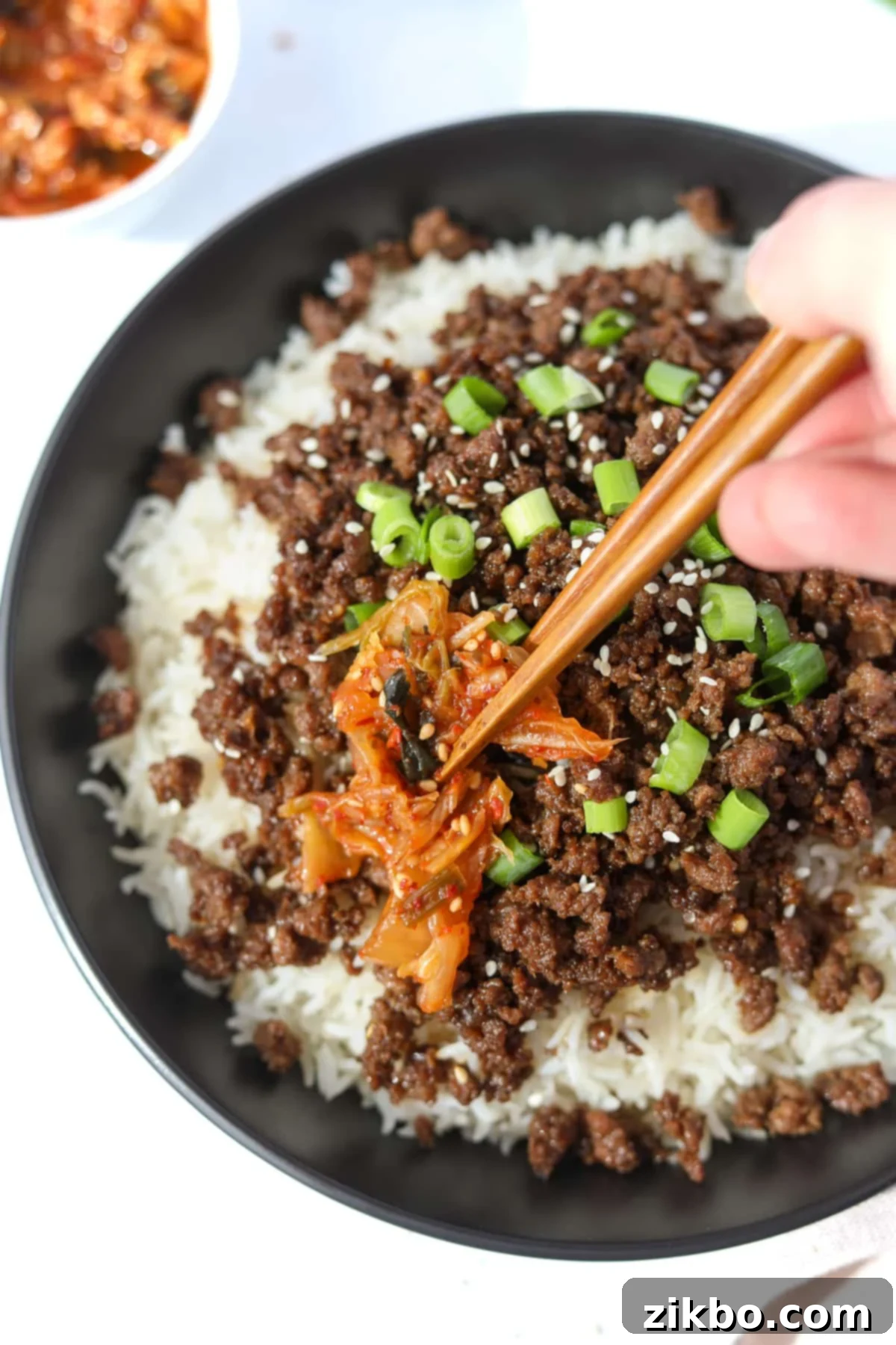 A hand placing a piece of vibrant red kimchi next to a portion of Korean ground beef and rice in a bowl.
