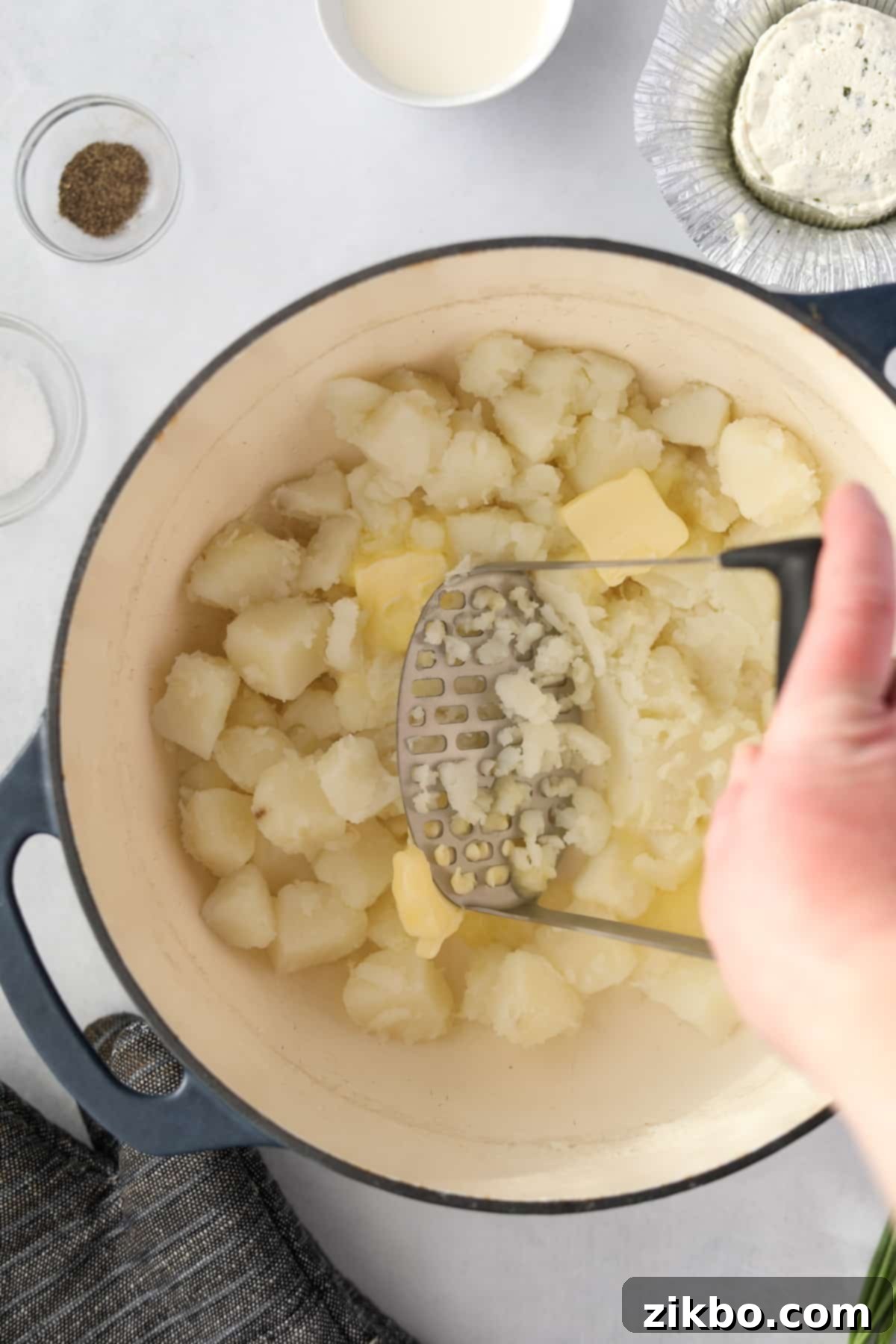 A potato masher actively mashing softened potatoes and butter in a Dutch oven, creating a smooth texture.