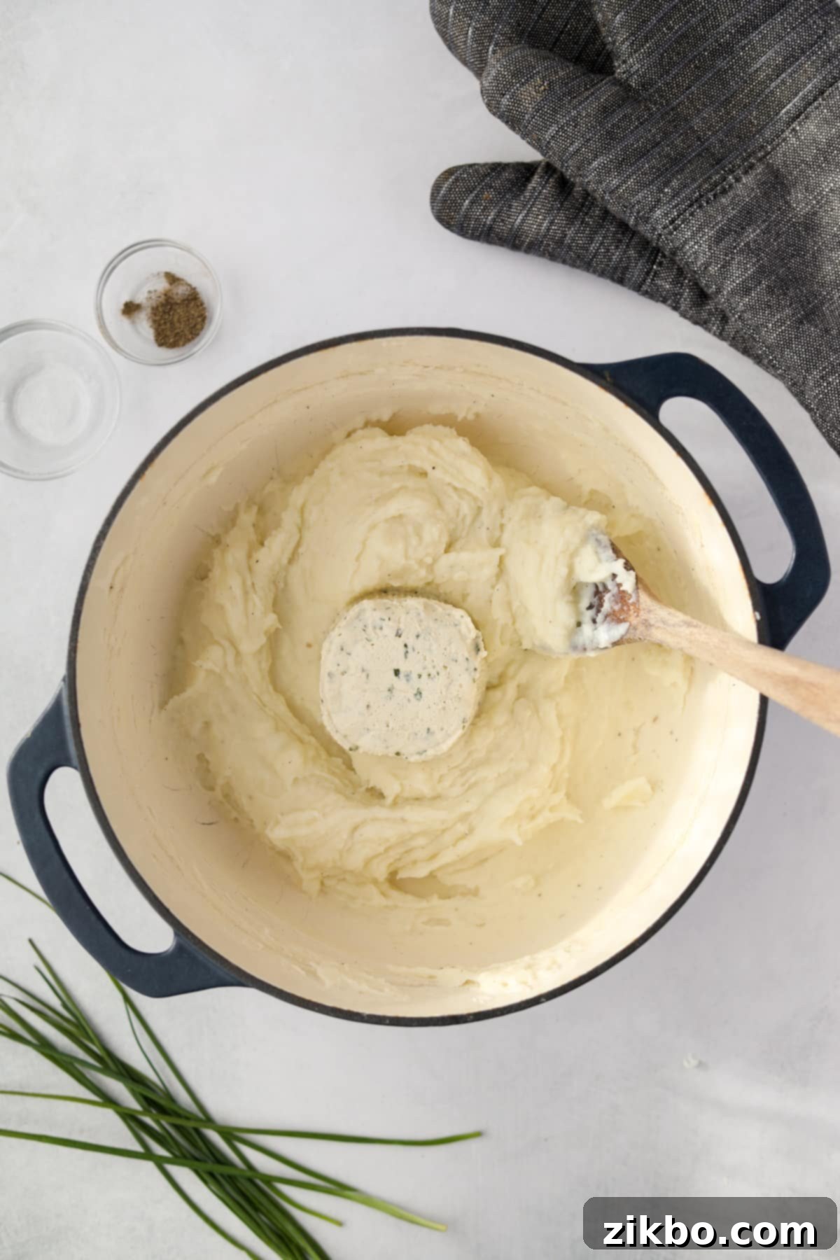 A Dutch oven filled with mashed potatoes and a full round of Boursin cheese, broken into pieces, ready to be incorporated.